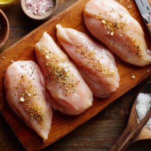 Raw chicken breasts seasoned with salt, pepper, and herbs on a wooden cutting board, ready for cooking. Bowls of pink Himalayan salt, sea salt, and olive oil are visible nearby.