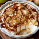 Homemadecaramel apple cheesecake dip in a white dish topped with sliced apples, graham cracker crumbs, and drizzled caramel sauce, ready to serve for dessert.