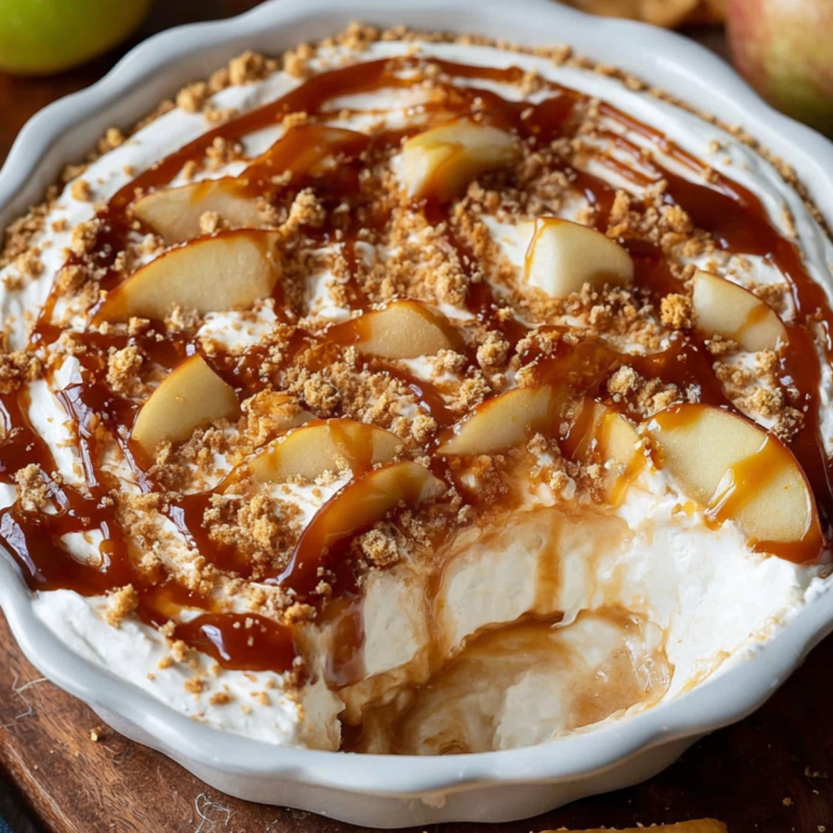 Homemadecaramel apple cheesecake dip in a white dish topped with sliced apples, graham cracker crumbs, and drizzled caramel sauce, ready to serve for dessert.