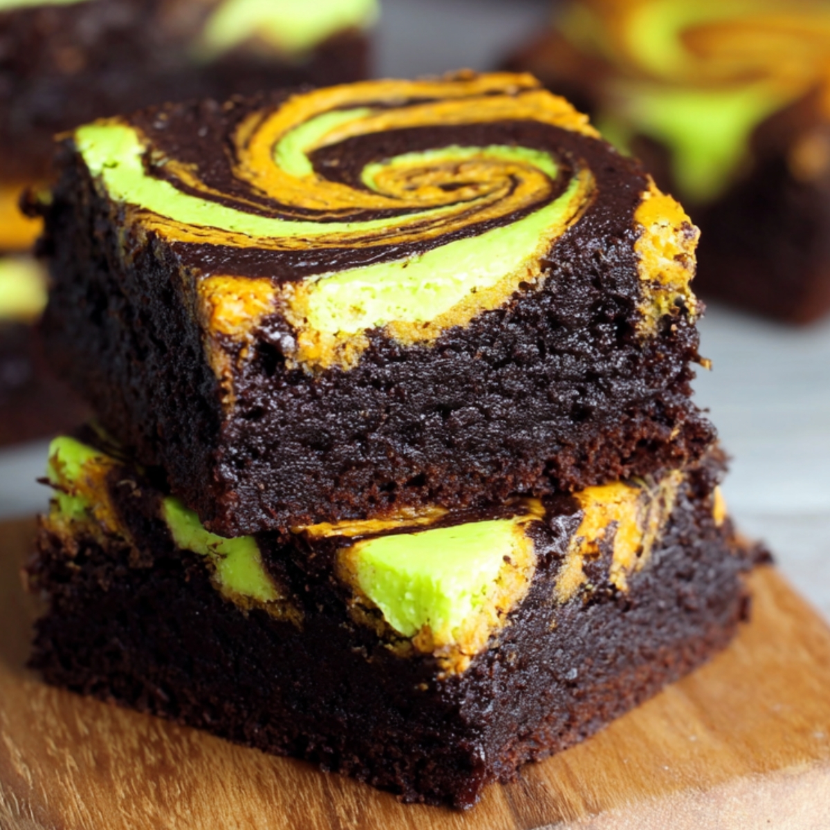 Stack of Halloween swirl cream cheese brownies with orange and green swirled tops over rich chocolate brownie layers, served on a wooden board.