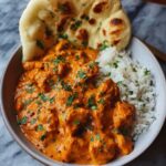 “Rich butter chicken served in a bowl with fluffy white rice and naan bread, topped with fresh cilantro.”