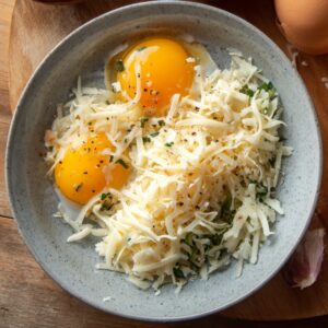 Two raw eggs in a gray ceramic bowl mixed with shredded mozzarella and herbs, ready to be whisked for cheesy pizza eggs.