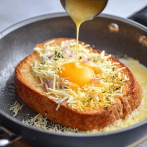 Golden toast in a skillet topped with shredded cheese and a cracked egg in the center, as creamy beaten egg is poured over for a melty, cheesy breakfast toast.