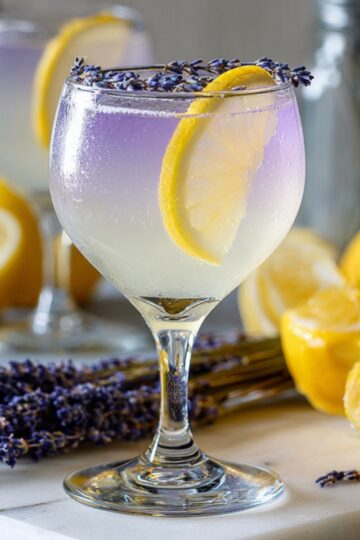 lavender limoncello spritz with a soft purple gradient, garnished with lemon slice and lavender buds on the rim, surrounded by fresh lemons and lavender flowers.
