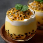 A glass dessert layered with yellow passion fruit mousse , white topping, and passion fruit pulp with seeds on top, garnished with a fresh mint leaf.