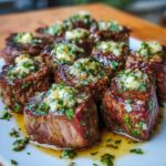 Cooked steak bites with garlic butter arranged on a white plate, each topped with a spoonful of garlic herb butter, with melted juices pooling at the bottom.