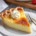 A slice of buttermilk pie custard pie with a caramelized top, served on a white plate and topped with whipped cream, with a fork beside it.