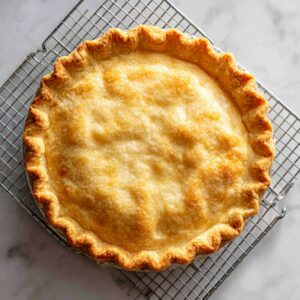 A whole baked pie with a golden, flaky crust, cooling on a metal wire rack over a marble surface.