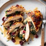 Slices of cranberry and spinach Christmas stuffed chicken breasts on a plate, with melted cheese and sauce, garnished with cranberries and herbs, served with a fork.