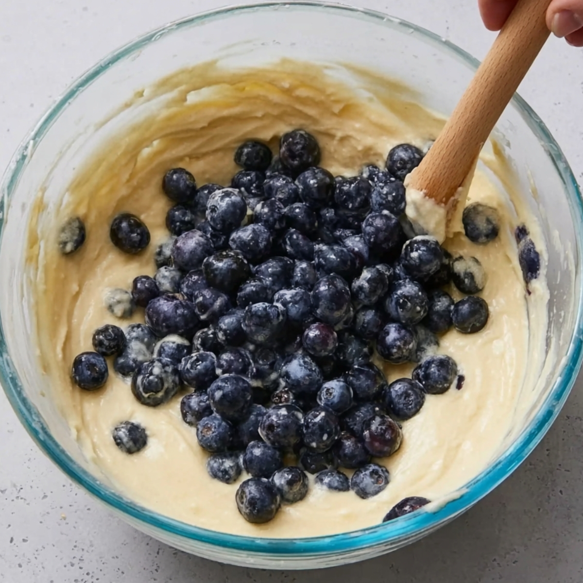 Folding fresh blueberries into cake batter in a glass mixing bowl with a wooden spoon
