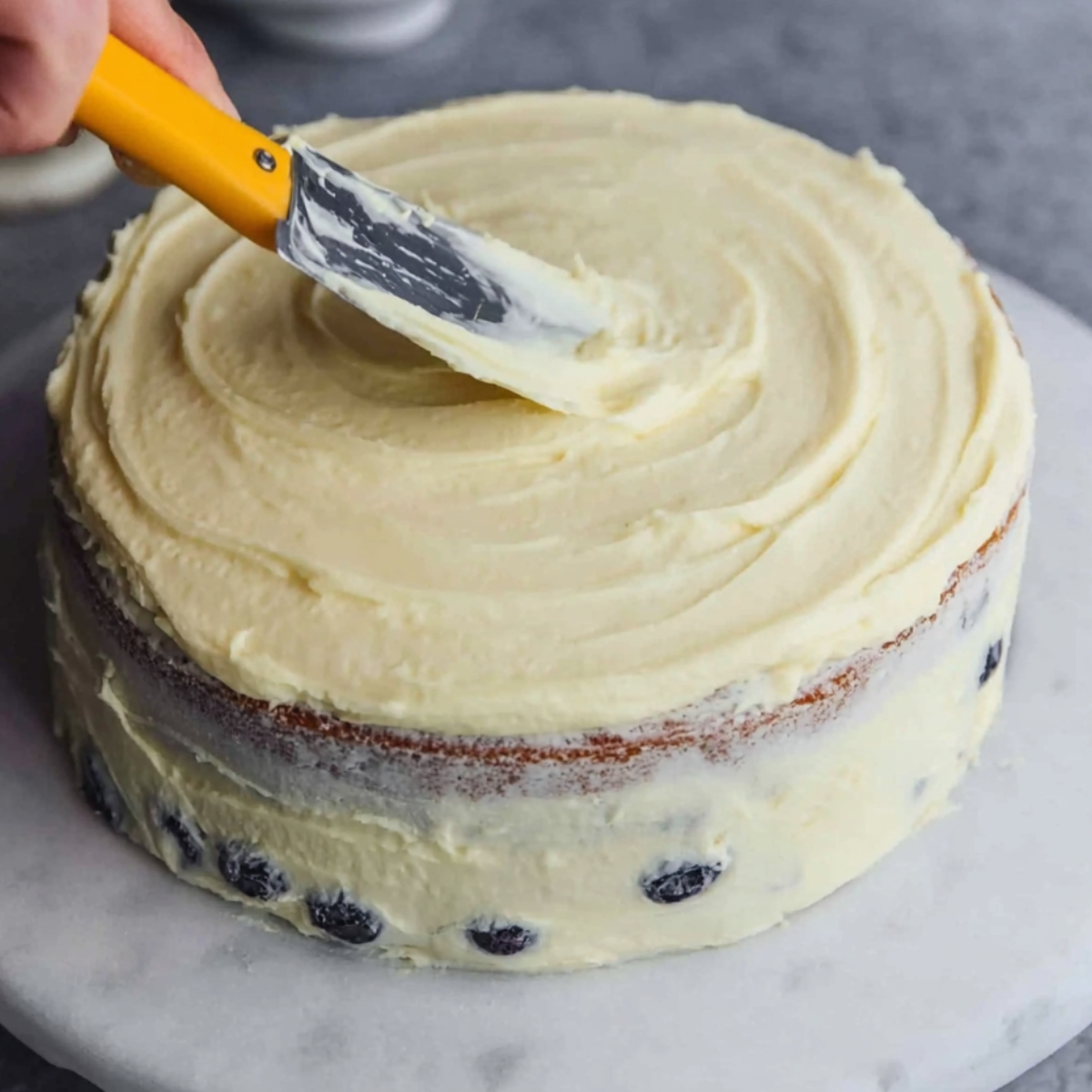 Spreading a layer of frosting on top of a lemon blueberry cake with a spatula.