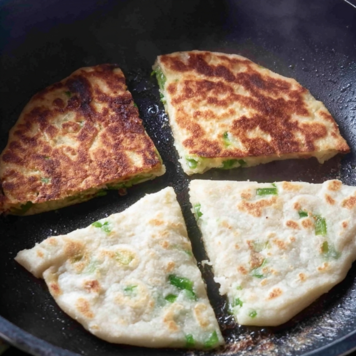 Triangular scallion flatbreads frying in a skillet, some golden-brown and some still pale.