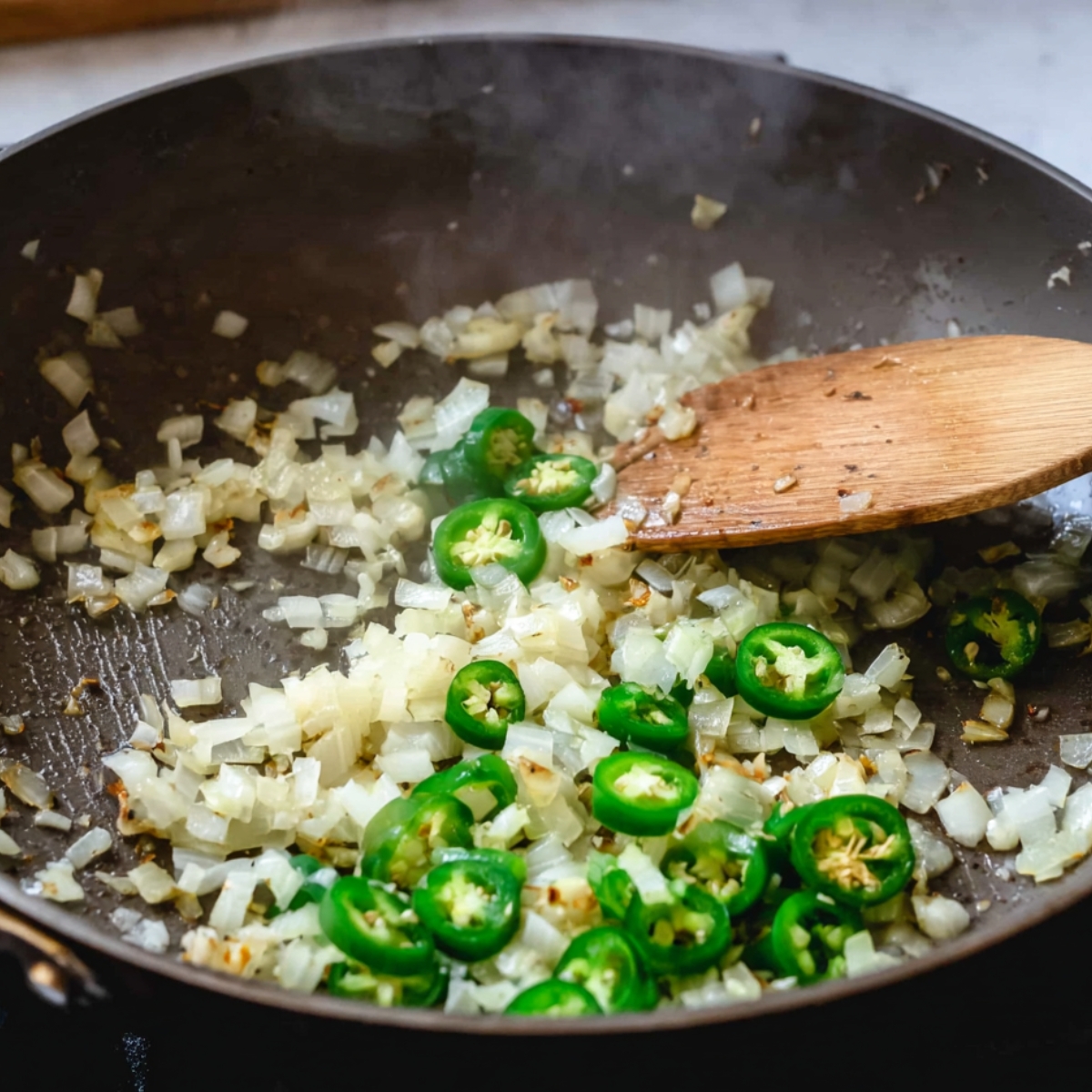 The image shows a pan with sautéed onions and sliced jalapeños, with a wooden spoon stirring the mixture, creating a flavorful base for a dish.