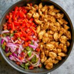 Overhead view of a skillet with seasoned chicken, red bell peppers, onions, and green peppers cooking side by side.