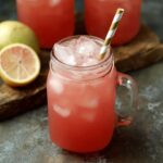 "Glass mason jar filled with iced guava white tea lemonade, garnished with a striped straw and a slice of lemon in the background."