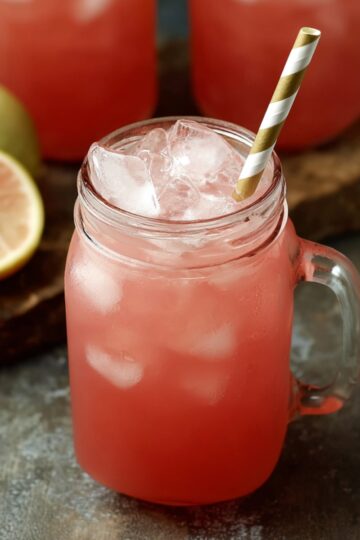 "Glass mason jar filled with iced guava white tea lemonade, garnished with a striped straw and a slice of lemon in the background."