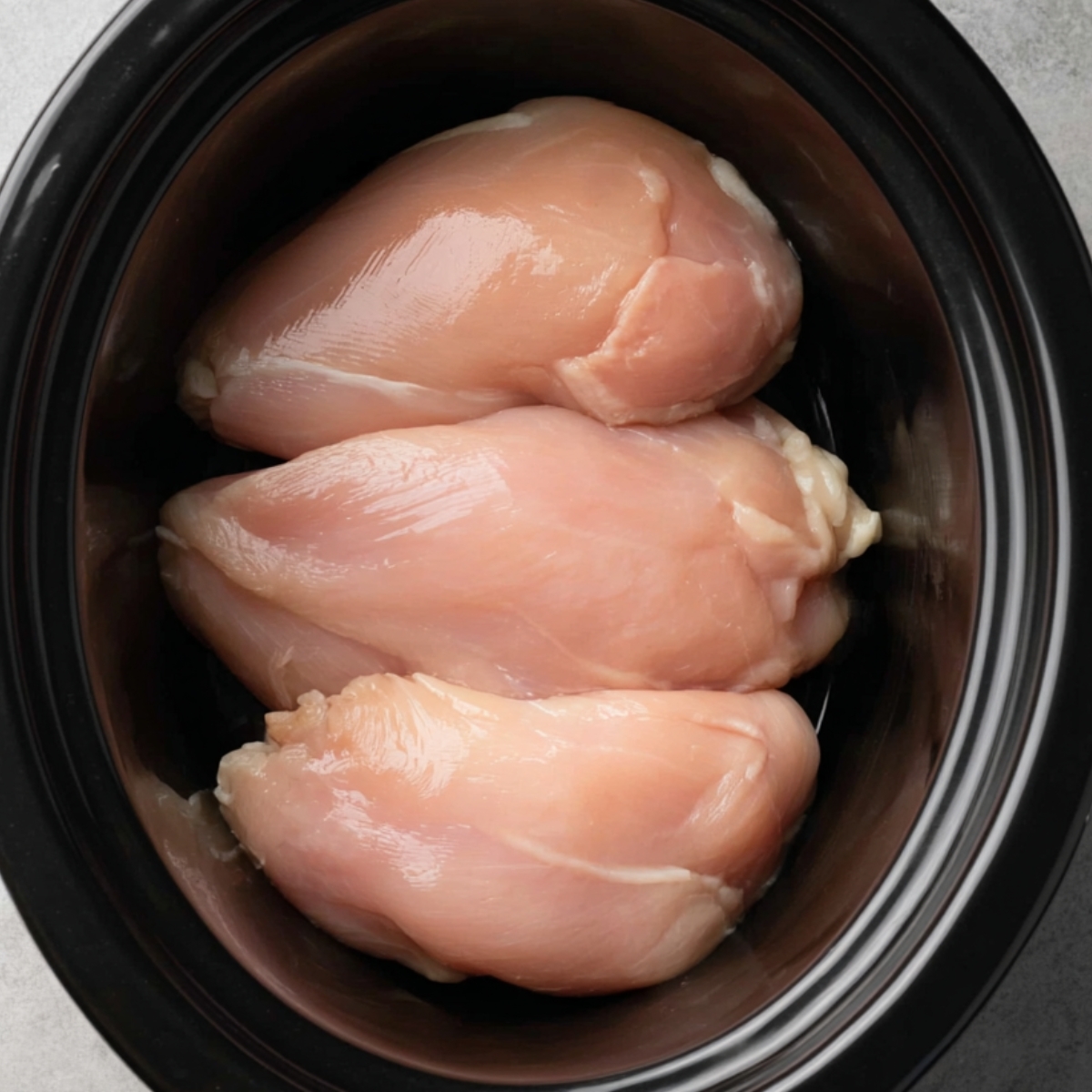 Three raw chicken breasts placed in the base of a slow cooker.