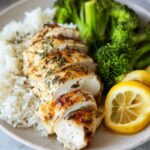 Sliced baked lemon chicken served with rice, broccoli, and lemon wedges.