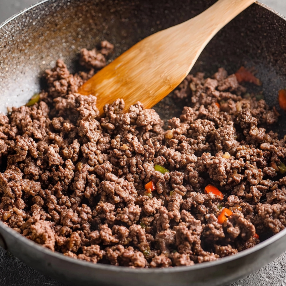 Ground beef being browned in a skillet with a wooden spoon, with some vegetables mixed in.