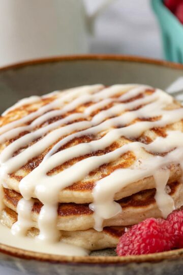 A stack of Cinnamon Roll Pancakes drizzled with icing and served with fresh raspberries.