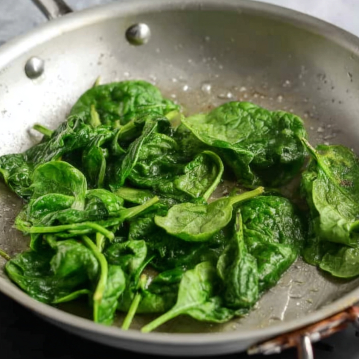 Fresh spinach leaves cooking in a silver skillet with a bit of oil.