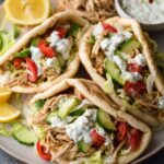 Three Crockpot Greek Chicken Gyros filled with shredded chicken, lettuce, cucumber, tomato, and topped with yogurt sauce, served with lemon wedges.