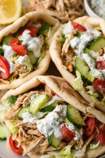 Three Crockpot Greek Chicken Gyros filled with shredded chicken, lettuce, cucumber, tomato, and topped with yogurt sauce, served with lemon wedges.
