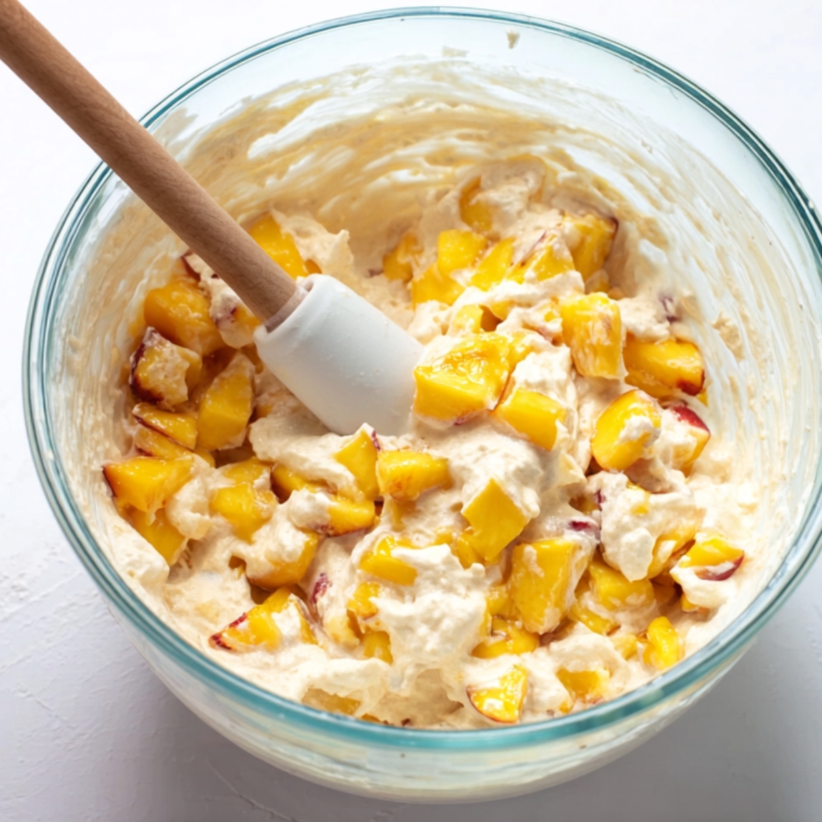 Fresh diced peaches folded into creamy cake batter in a glass bowl.
