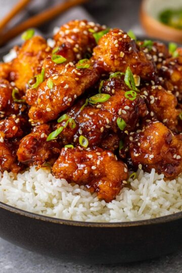 A bowl of General Tso Chicken served over rice, garnished with green onions and sesame seeds.