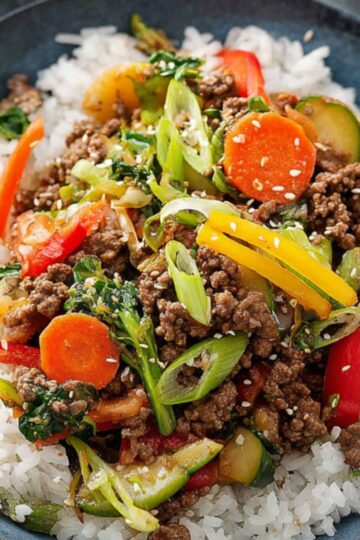 A bowl of white rice topped with Ground Beef Stir Fry, mixed with colorful vegetables like carrots, red peppers, and green onions, garnished with sesame seeds.