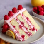 A slice of lemon raspberry cake with layers of fluffy cake, raspberry filling, and pink frosting, topped with fresh raspberries and a lemon slice.