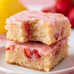 Two strawberry lemon blondies stacked on top of each other, drizzled with a pink glaze and a bite taken out, revealing the soft interior.