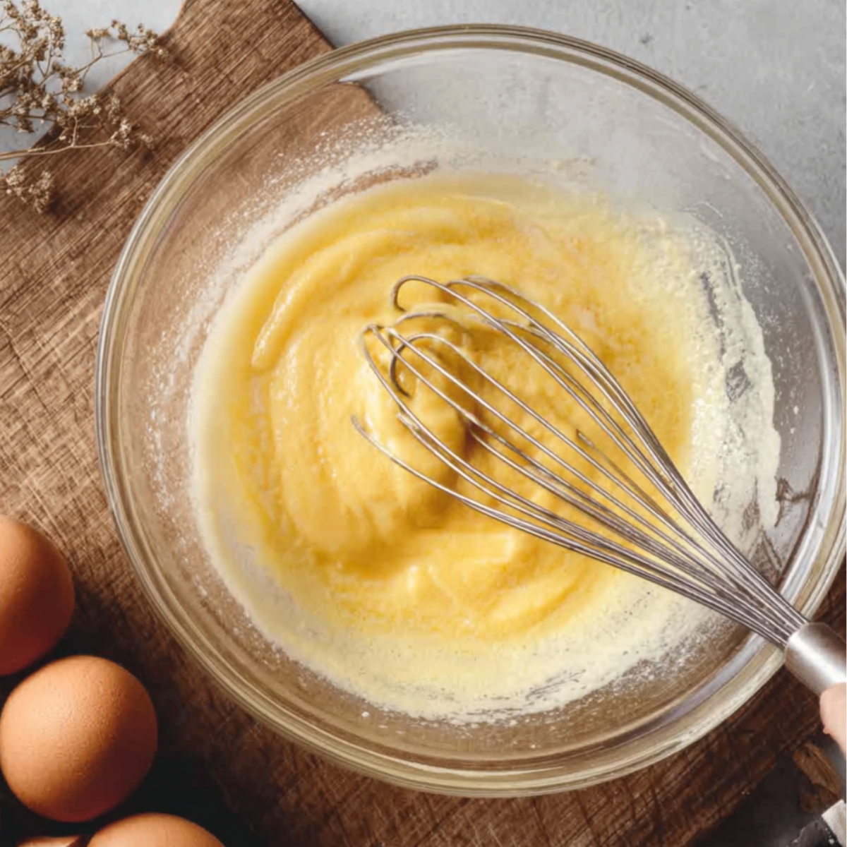 A person whisking eggs in a glass bowl, surrounded by fresh eggs on the table.