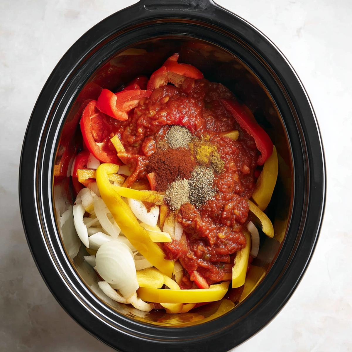 Top-down view of a black slow cooker filled with sliced red and yellow bell peppers and white onions, ready for cooking. Clean white countertop background."Top-down view of a black slow cooker filled with sliced red and yellow bell peppers and white onions, ready for cooking. Clean white countertop background.