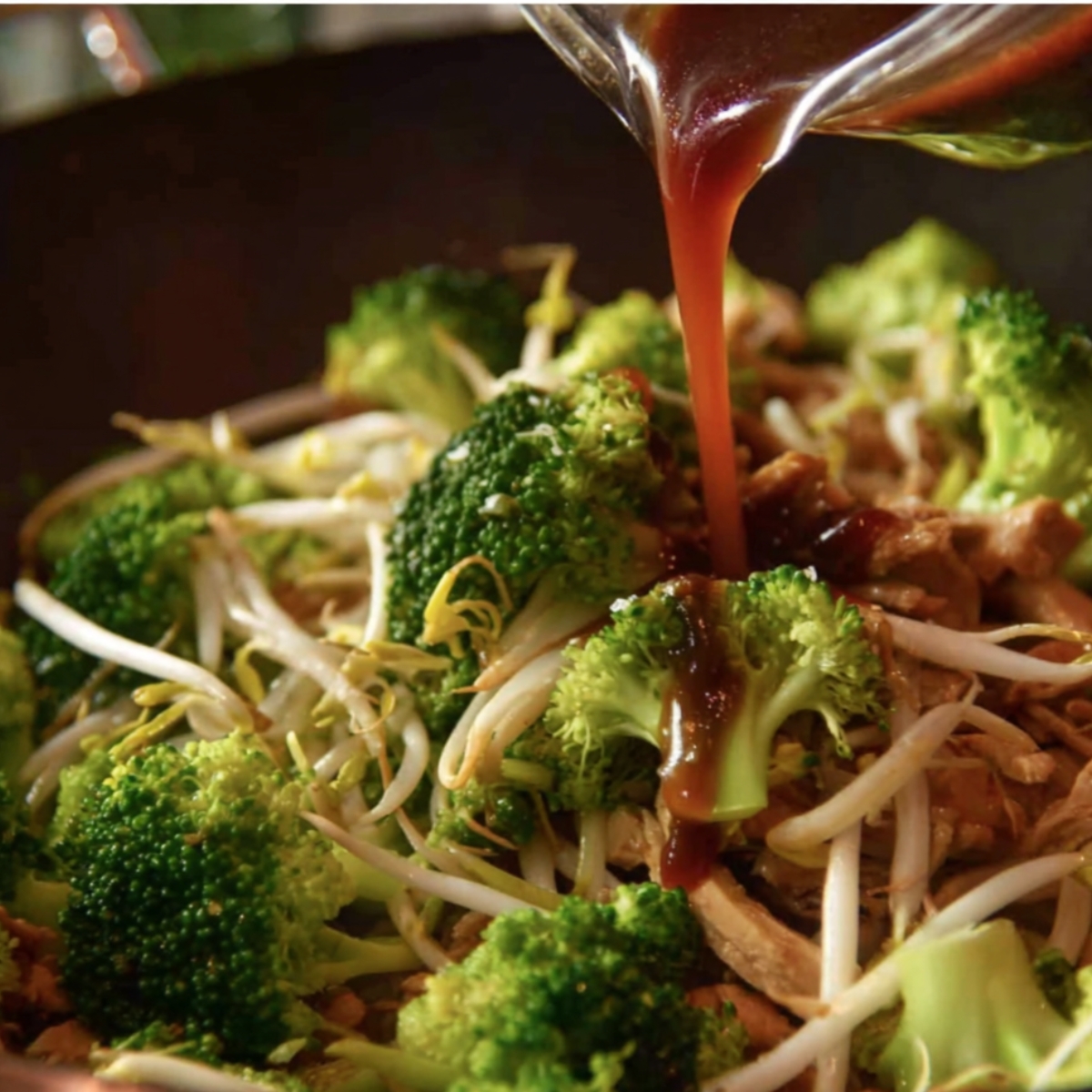 Close-up of broccoli and bean sprouts in a wok with brown sauce being poured over them, capturing the vibrant green vegetables and glossy sauce in mid-pour."