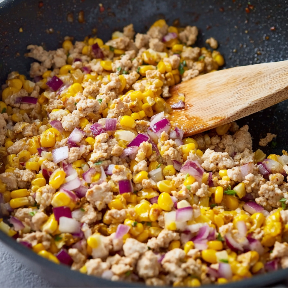 Close-up of ground chicken cooking in a skillet with sweet corn and diced red onions, stirred with a wooden spatula, showing colorful fresh ingredients and textures.