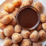 White plate filled with golden air-fried churro bites coated in cinnamon sugar, arranged around a small glass bowl of chocolate dipping sauce.