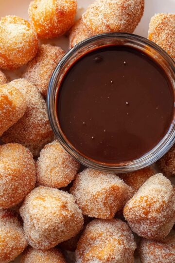 White plate filled with golden air-fried churro bites coated in cinnamon sugar, arranged around a small glass bowl of chocolate dipping sauce.