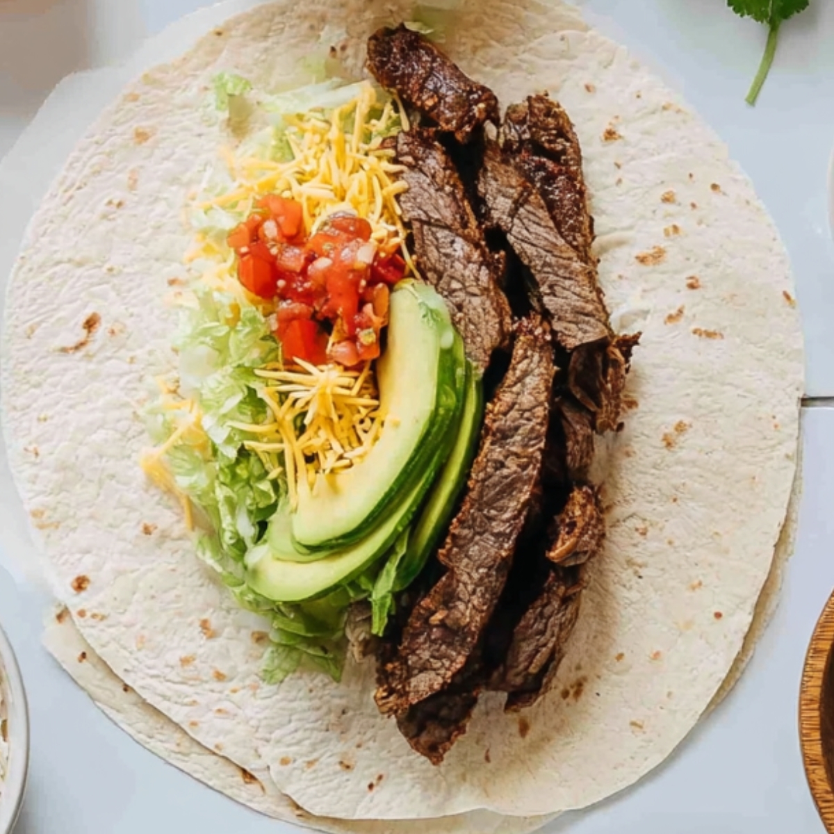 Top-down view of a large flour tortilla filled with sliced grilled steak, shredded cheese, sliced avocado, chopped lettuce, and diced tomatoes, ready to be rolled into a burrito.