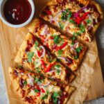 Cooked rectangular BBQ chicken flatbreads on a wooden cutting board, sliced into three pieces each. The flatbreads are golden brown and topped with melted mozzarella cheese, shredded BBQ chicken, red bell pepper slices, red onion strips, and fresh chopped cilantro. A small bowl of extra BBQ sauce is placed nearby, enhancing the presentation.