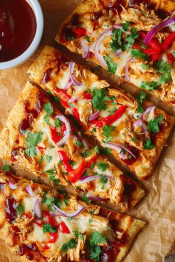 Cooked rectangular BBQ chicken flatbreads on a wooden cutting board, sliced into three pieces each. The flatbreads are golden brown and topped with melted mozzarella cheese, shredded BBQ chicken, red bell pepper slices, red onion strips, and fresh chopped cilantro. A small bowl of extra BBQ sauce is placed nearby, enhancing the presentation.