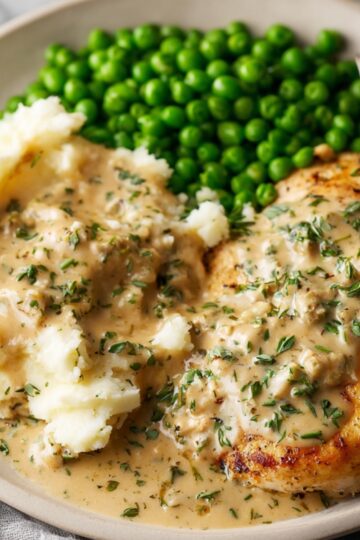 Plate of Boursin chicken with creamy herb sauce, served with mashed potatoes topped with sauce and bright green peas, fork on the side, home-cooked comfort food.
