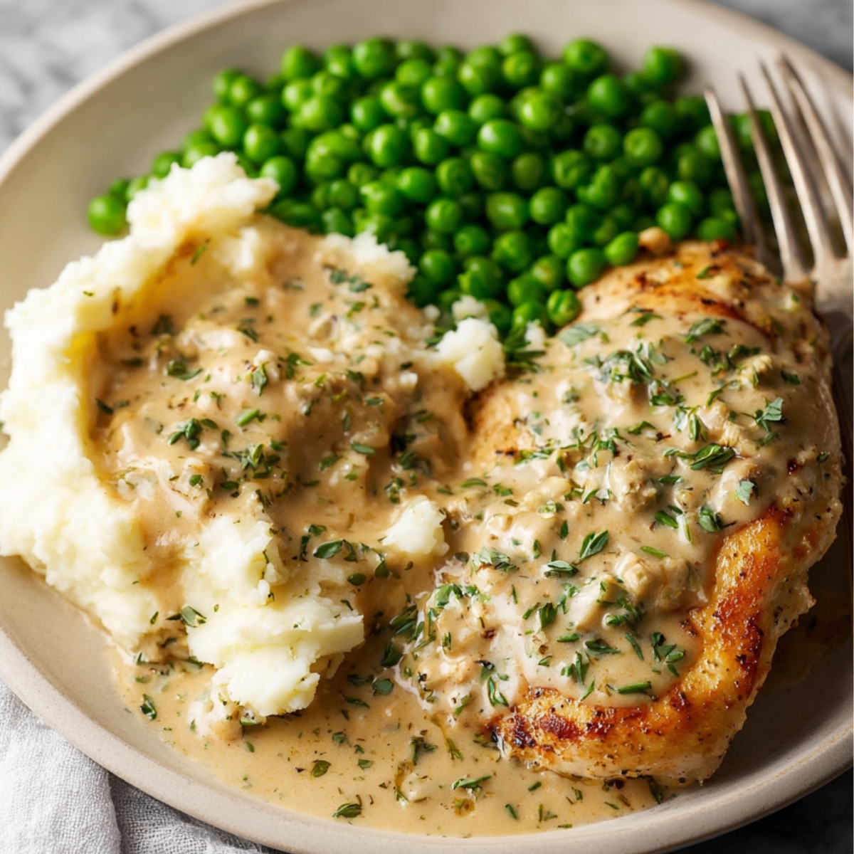 Plate of Boursin chicken with creamy herb sauce, served with mashed potatoes topped with sauce and bright green peas, fork on the side, home-cooked comfort food.