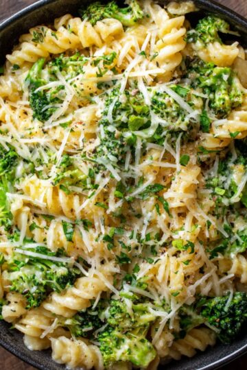 Cooked Broccoli Cheese Pasta fusilli pasta in a black skillet, mixed with melted cheese, garnished with grated Parmesan and chopped herbs, creamy and cheesy texture, top-down view, ready to serve as a comfort food dish.