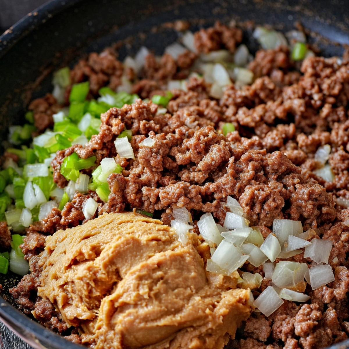 A skillet containing cooked ground beef with onions, green bell peppers, and a scoop of refried beans, prepared as the filling for chimichangas.