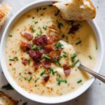 White bowl filled with creamy Cajun potato soup, garnished with crispy bacon pieces, chopped chives, and paprika, with a spoon and a piece of bread resting on the side.