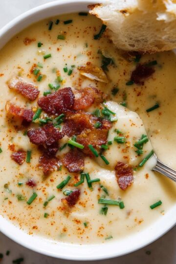 White bowl filled with creamy Cajun potato soup, garnished with crispy bacon pieces, chopped chives, and paprika, with a spoon and a piece of bread resting on the side.