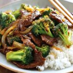 Chinese Beef and Broccoli stir-fry served over steamed white rice in a bowl, with tender beef slices coated in a glossy brown sauce and vibrant green broccoli, garnished with sautéed onions and chopsticks resting on the plate.
