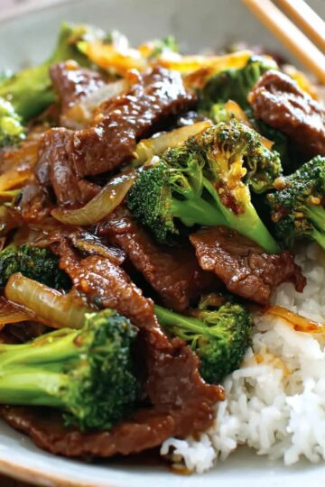 Chinese Beef and Broccoli stir-fry served over steamed white rice in a bowl, with tender beef slices coated in a glossy brown sauce and vibrant green broccoli, garnished with sautéed onions and chopsticks resting on the plate.