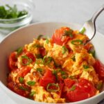 A bowl of Chinese Tomato Egg Stir-Fry over white rice, with soft scrambled eggs, juicy red tomato pieces, and garnished with sliced green onions, with a fork lifting a portion of the dish.